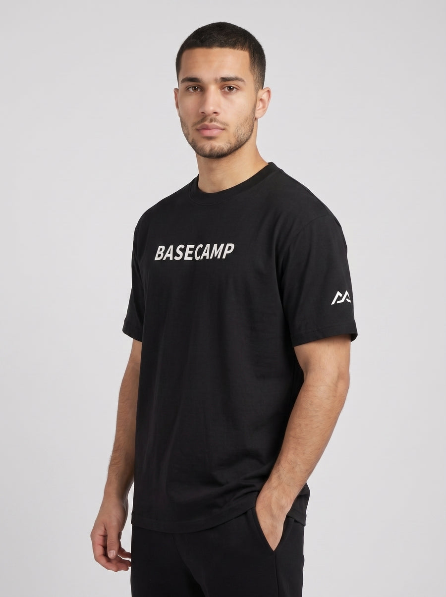 Model wearing the black BASECAMP Heavyweight Tee, three-quarter view with hand in pocket, BASECAMP wordmark across the chest and the Marko two-peak mark on the sleeve