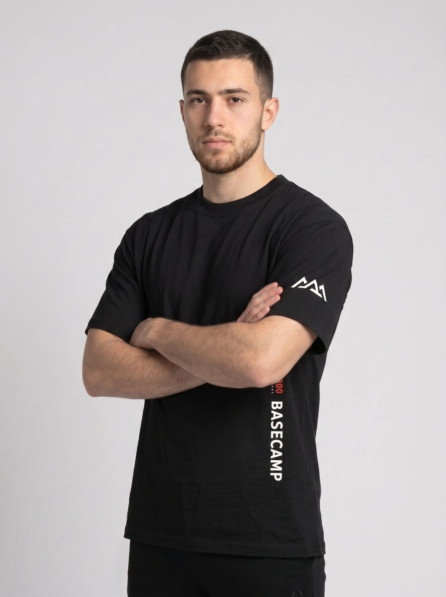 Model wearing the black 300 Heavyweight Tee, arms crossed, 001/300 First Ascent and BASECAMP printed vertically at the hem and the Marko two-peak mark on the sleeve